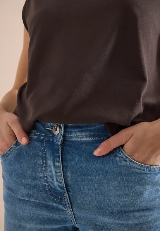 Een persoon draagt een donkerbruin shirt en blauwe jeans, handen in de zakken.