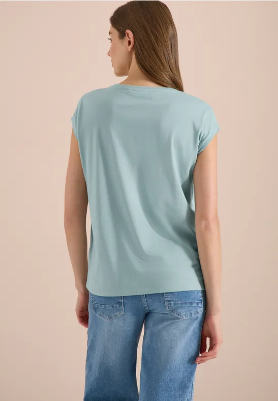 Frau von hinten in hellblauem lockerem T-Shirt und blauen Jeans vor neutralem Hintergrund.