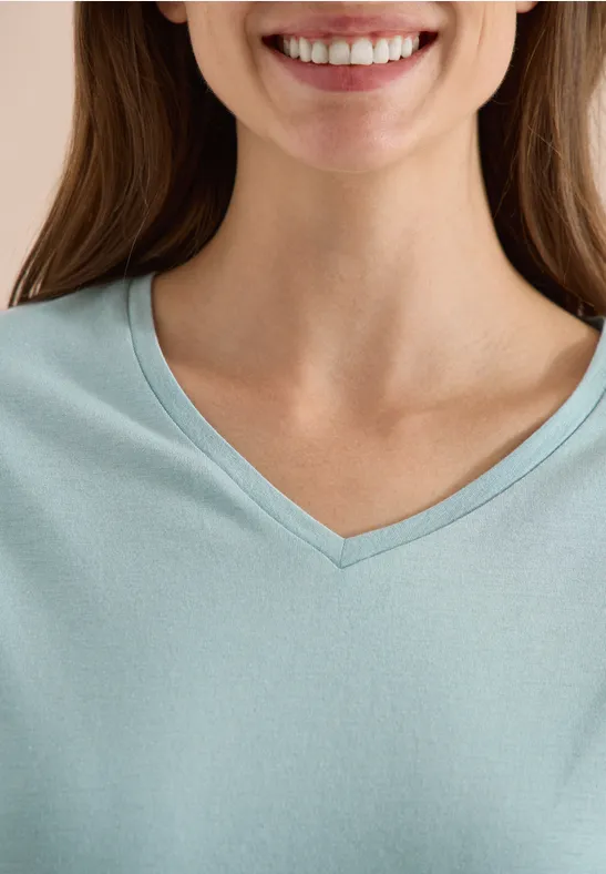 Lächelnde Frau im hellblauen V-Ausschnitt-Shirt, nur Hals und Brustansatz sichtbar.