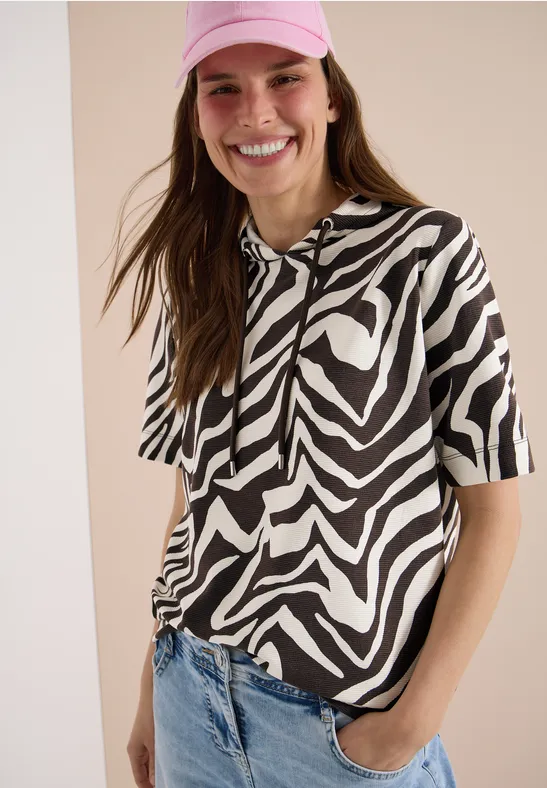 Frau vor hellem Hintergrund trägt kurzärmeliges Zebra-Kapuzen-Shirt in Schwarz-Weiß und blaue Jeans.