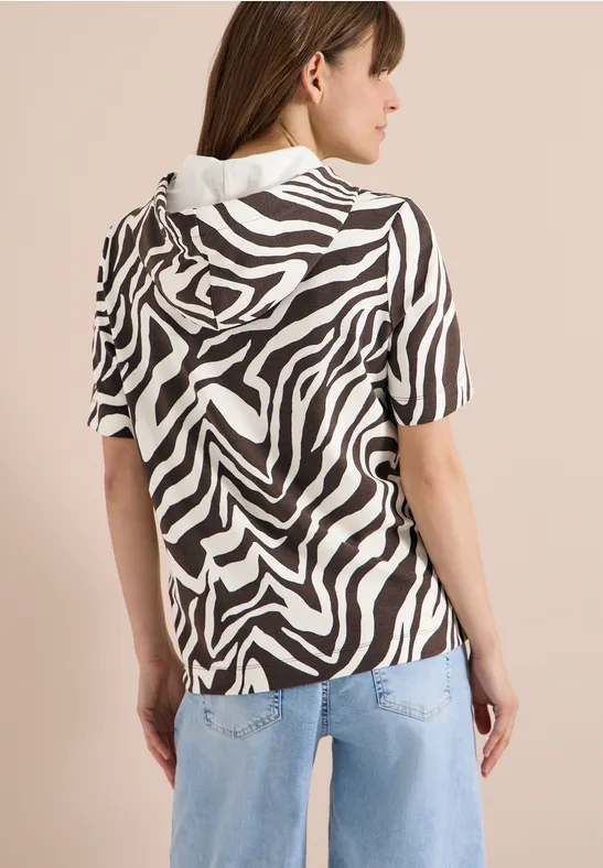 Frau von hinten in kurzärmeliger Zebra-Kapuzenbluse und hellblauen Jeans vor neutralem Hintergrund.