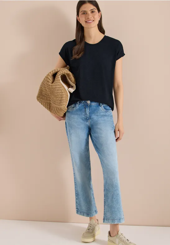 Vrouw voor beige muur met zwart shirt, lichtblauwe jeans, sneakers en gevlochten strooien tas.