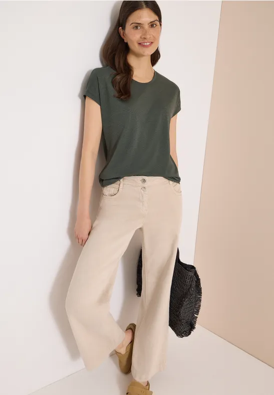 Een vrouw leunt tegen een witte muur, draagt een donkergroen T-shirt, licht wijde jeans, gouden schoenen en een zwarte tas.