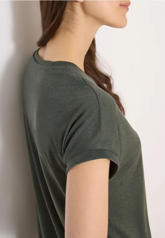 Jonge vrouw in zijaanzicht, leunt tegen een witte muur, draagt een donkergroen kort T-shirt, kijkt voor zich