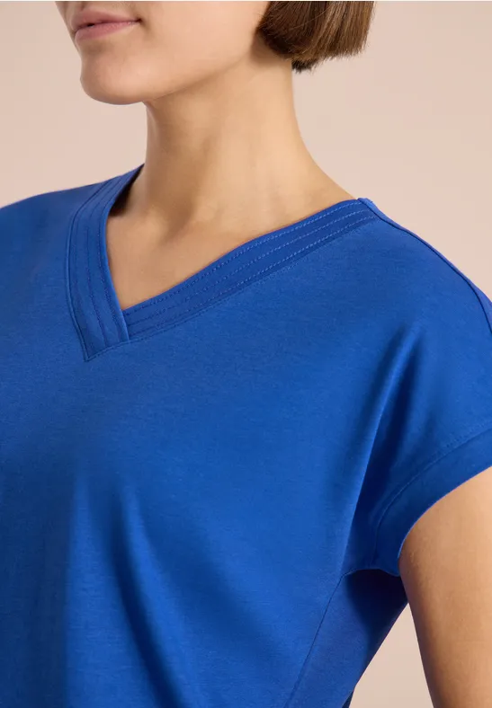 Nahaufnahme einer Person in royalblauem V-Ausschnitt-Shirt vor neutralem Hintergrund.