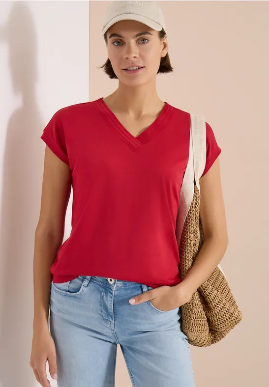 Frau trägt rotes V-Shirt, helle Jeans und eine Basttasche vor neutralem Hintergrund.