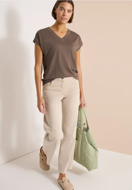 Frau in dunkelgrauem V-Ausschnitt-Shirt, hellen Schlaghosen, braunen Loafern und grüner Stepp-Tasche vor beiger Wand.