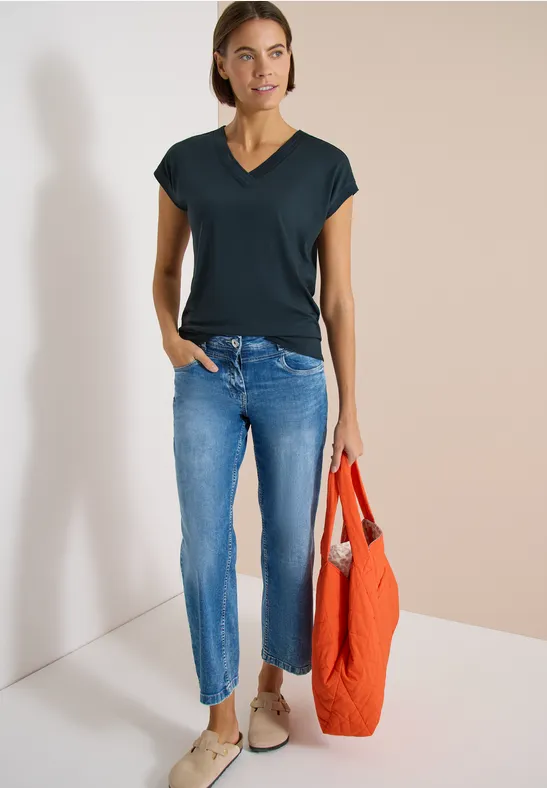 Person steht vor heller Wand, trägt schwarzes V-Shirt, blaue Jeans, beigen Schuhen und einen orangen Stoffbeutel.