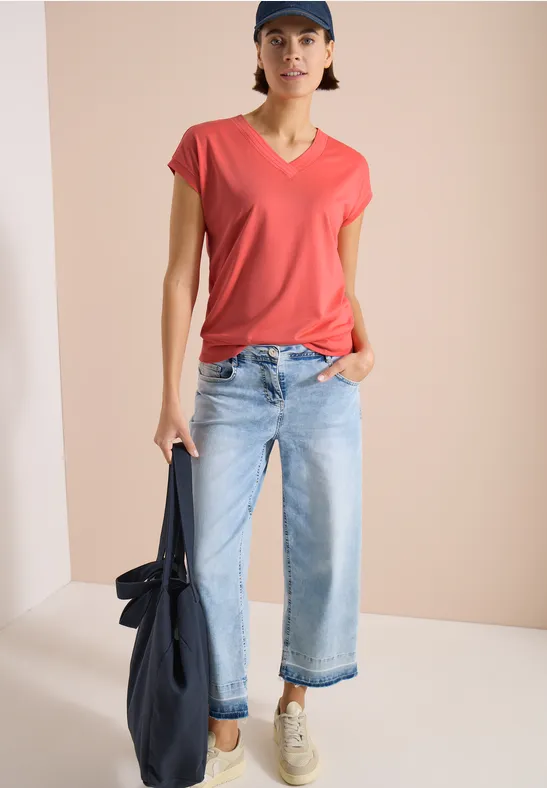 Person vor beigem Hintergrund trägt rotes T-Shirt, helle Jeans, Sneakers und hält eine Tasche.