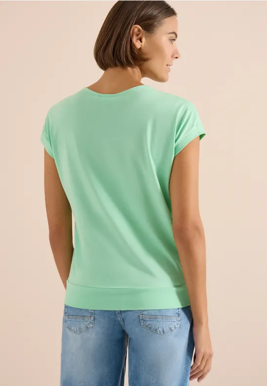 Rückansicht einer Person in mintgrünem T-Shirt und Jeans vor hellem Hintergrund.