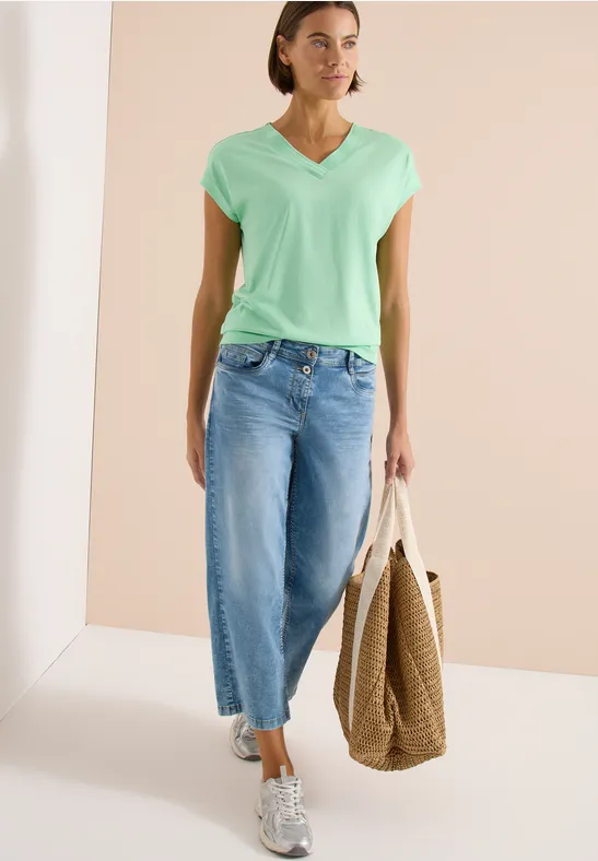Frau in mintfarbenem V-Shirt, weiten Culotte-Jeans, Sneakers und Bast-Shopper.