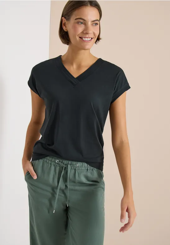 Vrouw in zwart V-hals T-shirt en groene joggingbroek, hand in de zak, tegen een neutrale achtergrond.