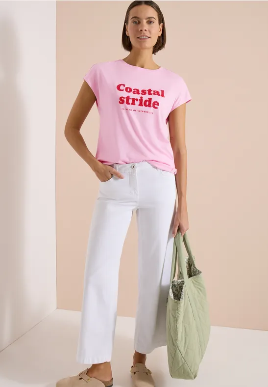 Vrouw draagt roze T-shirt met "Coastal Stride", witte wijde cropped jeans en groene gewatteerde shopper.