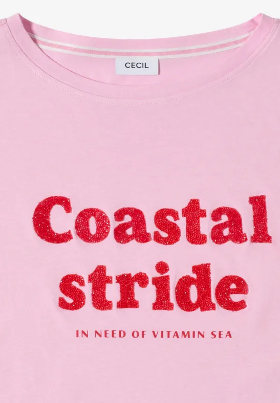 Een lichtroze T-shirt met rode glinsterende tekst 'Coastal stride.' en 'In need of vitamin sea'.