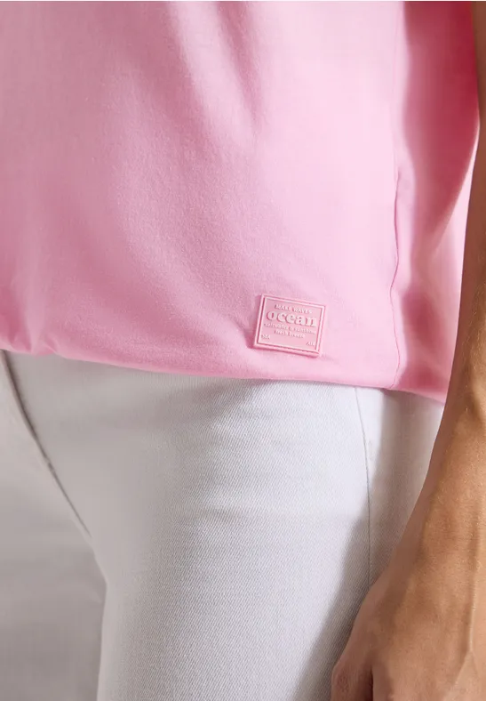 Een roze T-shirt met een klein label in de zoom, gedragen op een witte broek.