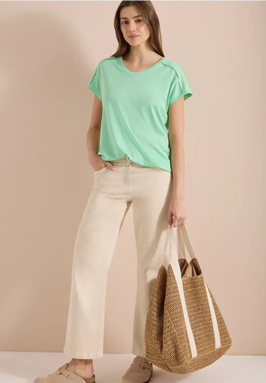 Eine Frau trägt ein mintgrünes T-Shirt, helle weite Hose und hält eine große Strohtasche.