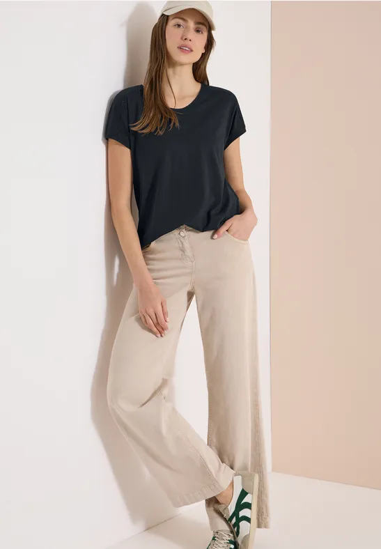 Frau lehnt entspannt an Wand in schwarzem T-Shirt und beigen, weiten Hosen.