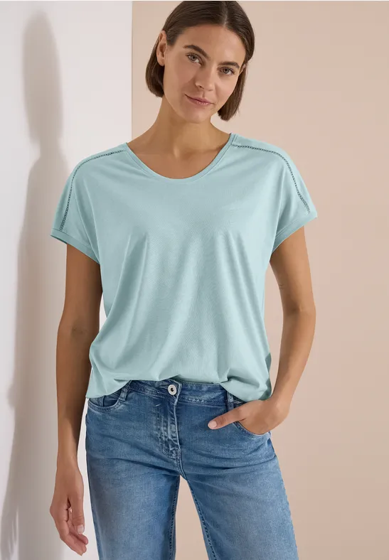 Frau in türkisfarbenem T-Shirt und Jeans lehnt entspannt vor hellem Hintergrund.