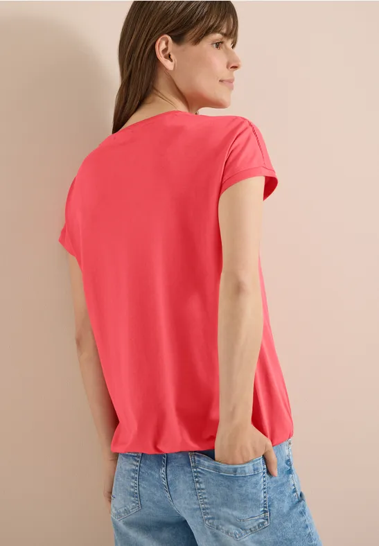 Rückansicht einer Frau in rotem T-Shirt und Jeans, eine Hand in der Hosentasche.
