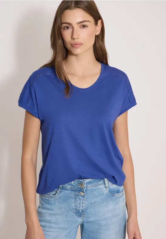 Vrouw draagt een blauw T-shirt en spijkerbroek, staand voor een neutrale achtergrond.
