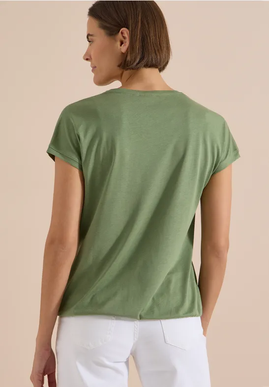 Eine Frau von hinten in grünem T-Shirt und weißer Hose vor schlichtem Hintergrund.