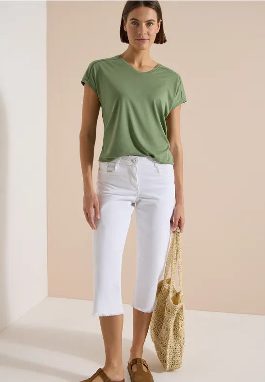 Frau trägt grünes T-Shirt, weiße Capri-Hosen und hält eine geflochtene Strohtragetasche.