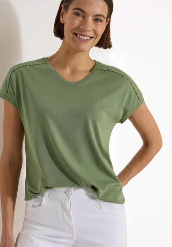 Eine Person trägt ein schlichtes grünes T-Shirt und weiße Jeans, Oberkörper sichtbar.