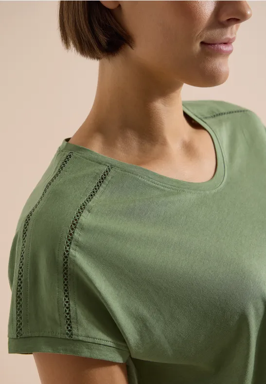 Nahaufnahme einer Frau in olivgrünem T-Shirt mit dekorativer Nahtverzierung an der Schulter.