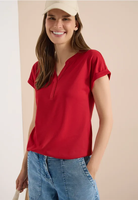Frau trägt rotes Kurzarmshirt mit V-Ausschnitt und blaue Jeans vor einfarbig hellem Hintergrund.