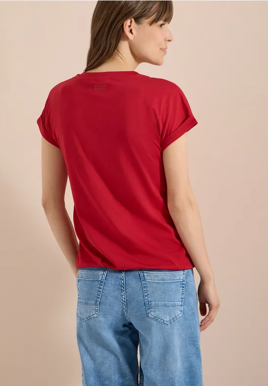 Frau steht Rücken zur Kamera und trägt rotes T-Shirt sowie blaue Jeans.