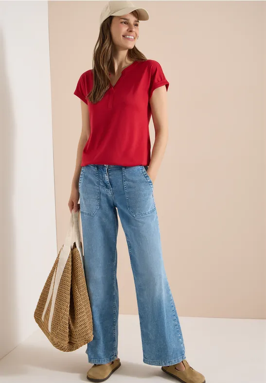 Frau in rotem T-Shirt, weiten blauen Jeans und geflochtener Strohtasche vor neutralem Hintergrund.