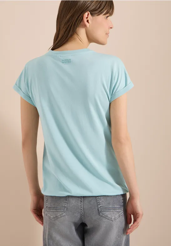 Frau von hinten in hellblauem T-Shirt und grauen Jeans vor beigem Hintergrund, Gesicht unscharf.