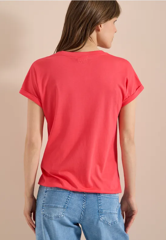 Frau von hinten in rotem T-Shirt und blauen Jeans vor beigem Hintergrund.
