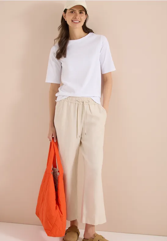 Vrouw in witte T-shirt en beige wijde broek met oranje tas, staande voor een beige achtergrond.