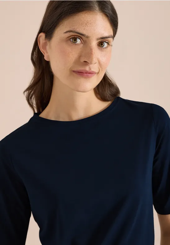 Persoon met een donkerblauw T-shirt met korte mouwen voor een lichte achtergrond, gezicht vervaagd.