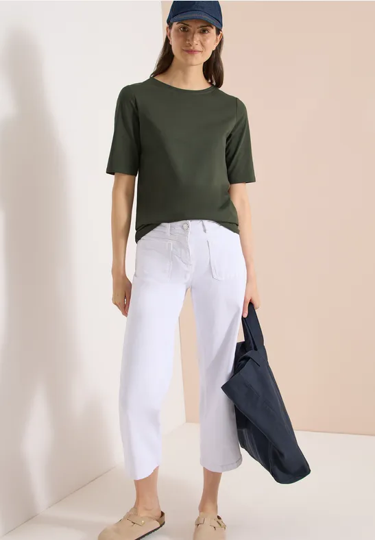 Vrouw in groen T-shirt met witte 7/8 wijdvallende broek, houdend een donkerblauwe jas in een minimalistische setting.