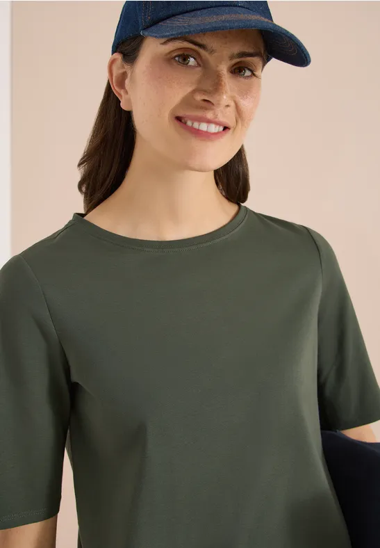 Persoon met olijfgroen t-shirt met halve mouwen en pet voor lichte achtergrond.