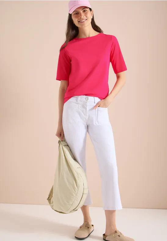 Vrouw in roze T-shirt en witte broek met een beige tas voor een beige muur.