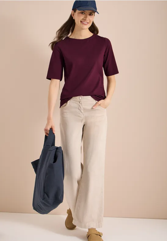 Vrouw in paars T-shirt en beige palazzo-broek met een donkerblauwe tas voor een eenvoudige achtergrond.