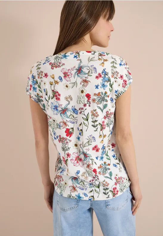 Vrouw van achteren in wit shirt met kleurrijke bloemenprint en blauwe jeans.