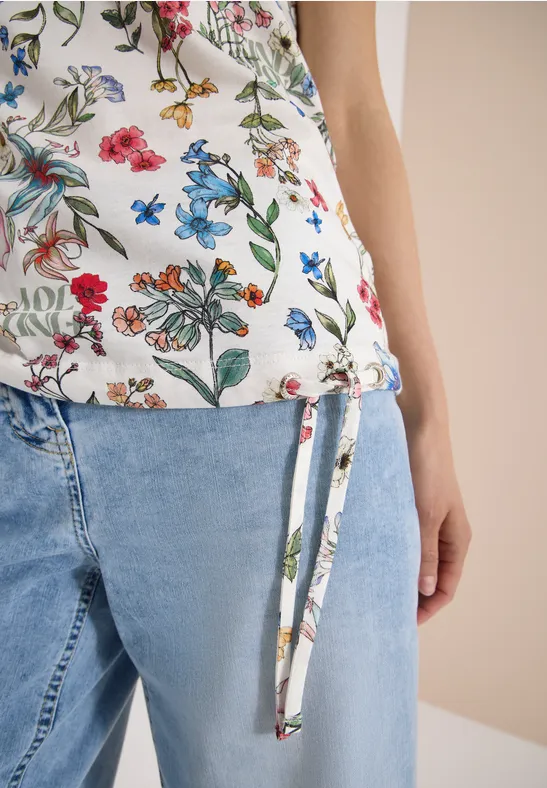 Close-up van een vrouw in een gebloemde top met zijstrik en lichtblauwe jeans.