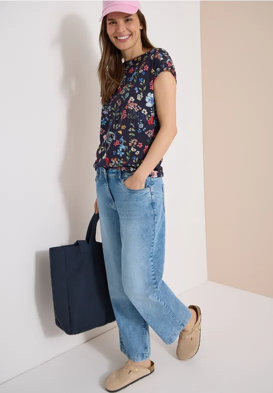 Persoon draagt een bloemen T-shirt, losse blauwe jeans, pantoffels en heeft een donkerblauwe tas.