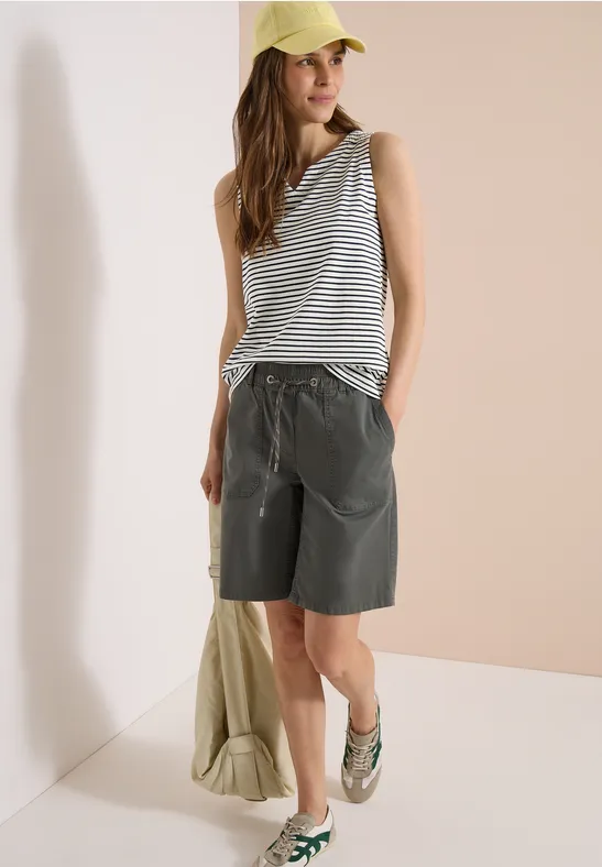 Person trägt gestreiftes Top, lockere Bermudashorts, Sandalen und Cap vor hellem Hintergrund.