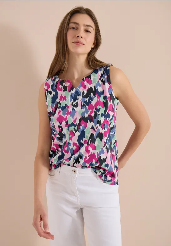 Vrouw draagt kleurrijke mouwloze top met abstract patroon en witte broek voor neutrale achtergrond.