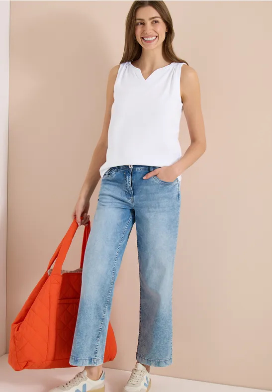 Frau in weißem Top und hellblauen Jeans mit großer orangefarbener Tasche vor neutralem Hintergrund.