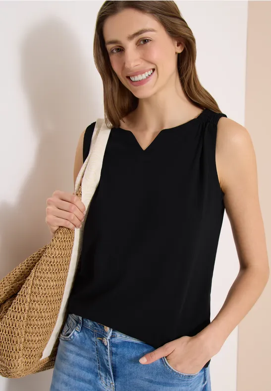 Frau in schwarzer ärmelloser Bluse trägt einen Korb­tasche und hat eine Hand in der Jeans­tasche.