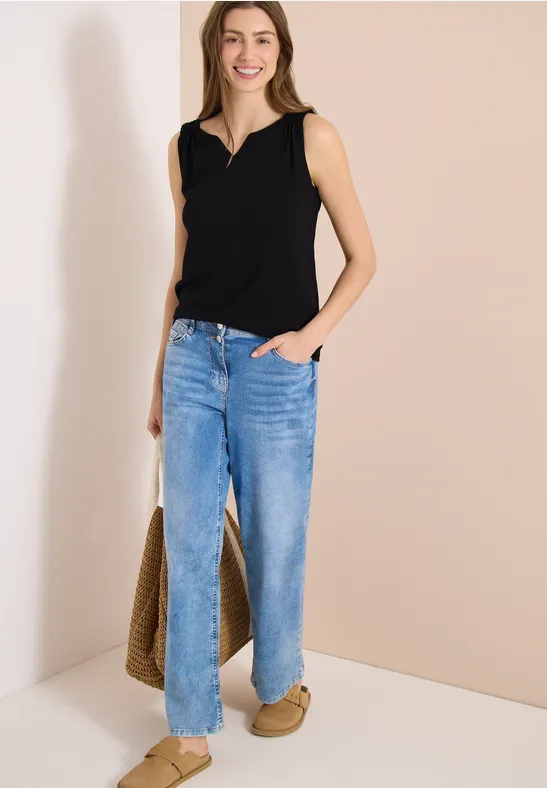 Frau in schwarzem Top und blauen Jeans steht vor hellem Hintergrund, trägt Korbtasche und Hausschuhe.