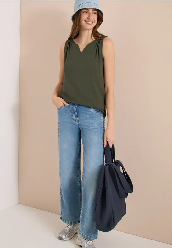 Eine Frau trägt eine ärmellose grüne Bluse, weite helle Jeans und hält eine dunkelblaue Tasche.