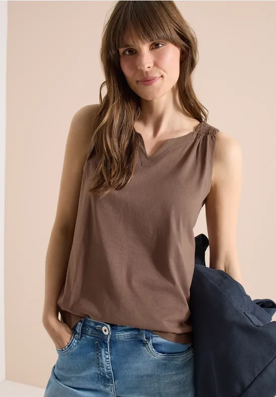 Frau in braunem, ärmellosem Top und Jeans, hält eine Jacke in der Hand.