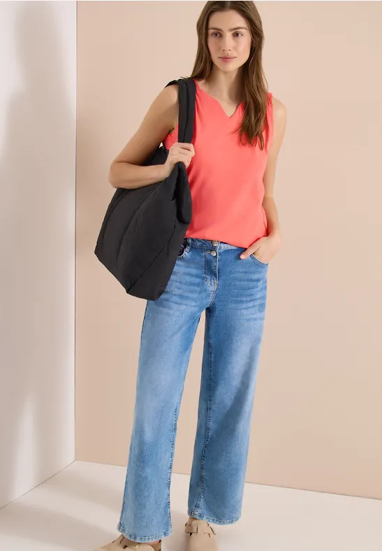 Vrouw met rood mouwloos topje, wijde blauwe jeans en grote zwarte tas voor een neutrale achtergrond.